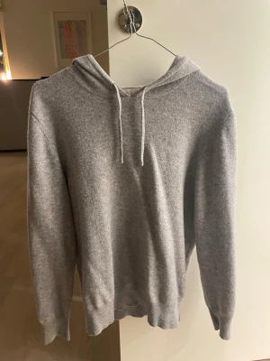 Sayless - Säljer en stilren grå hoodie från Sayless i mjuk ullmix. Hoodien har dragsko i huvan, ribbade muddar vid ärmslut och nederkant samt en relaxed passform. Perfekt för dig som gillar en clean och enkel look. Den har en liten slitning på den vänstra baksidan av armen. 