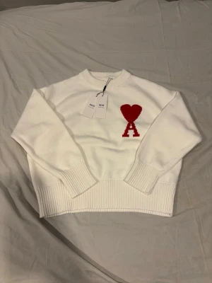 Vit stickad tröja från Ami Paris - Vit stickad tröja från Ami Paris med röd hjärtlogga och bokstaven A på bröstet. Tröjan har rund hals, ribbade muddar och nederkant. Perfekt för dig som gillar stilrena plagg med en twist.