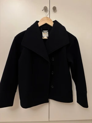 Mörk blå kappa med bred krage - Stilren mörkblå kort kappa med bred krage och tre knappar framtill. Klassisk rak passform och diskret design som funkar till många olika looks. Perfekt för dig som gillar minimalistisk stil och vill ha en kappa som känns både modern och tidlös.