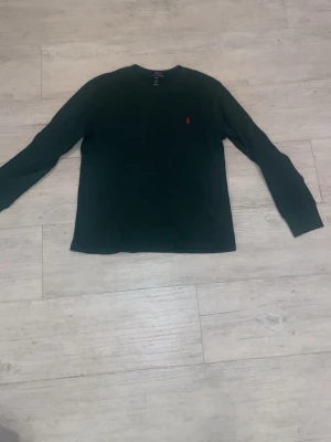 Mörkgrön långärmad tröja Polo Ralph Lauren - Mörkgrön långärmad tröja från Polo Ralph Lauren med rund halsringning och den klassiska röda logotypen broderad på bröstet. Tillverkad i ett mjukt, strukturerat material som känns skönt mot huden. Perfekt för en avslappnad och stilren look.