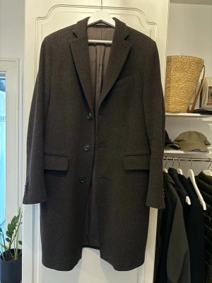 Mörkbrun ullrock från Oscar Jacobson - Stilren mörkbrun rock från Oscar Jacobson i klassisk rak modell. Rocken har tre knappar framtill, två fickor med lock och en bröstficka. Fodrad insida med innerficka och diskret logga. Perfekt för en snygg och tidlös look.