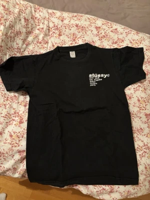 Svart Stüssy t-shirt - Säljer en svart t-shirt från Stüssy med tryck på bröstet och en stor jordgubbe på ryggen. Texten listar städer som New York, Los Angeles, Tokyo, London och Paris. Klassisk passform och rund hals. Perfekt för dig som gillar streetwear och coola prints.