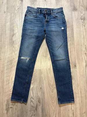Abercrombie & Fitch - Snygga blå jeans från Abercrombie & Fitch med slitningar på låret och knät. Jeansen har klassisk femficksdesign, dragkedjegylf och en tvättad look med ljusare partier. Perfekt för en avslappnad och trendig stil.