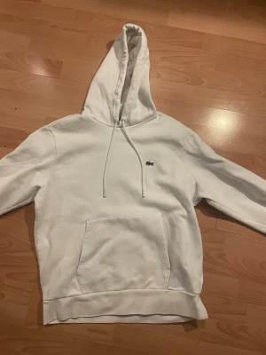 Lacoste hoodie - Säljer en clean vit hoodie från Lacoste i klassisk passform. Hoodien har dragsko i huvan, stor magficka och den ikoniska gröna krokodilloggan broderad på bröstet. Perfekt för dig som gillar stilrena plagg med premiumkänsla.