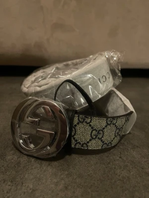 Gucci GG Supreme bälte med silverdetalj - Snyggt bälte från Gucci med klassiskt GG Supreme-mönster i beige och svart. Bältet har ett stort, blankt spänne i silver med GG-logga. Perfekt accessoar för att lyfta vilken outfit som helst.