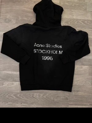 Svart hoodie Acne Studios Stockholm - Svart hoodie från Acne Studios med stor huva och tryck på ryggen där det står 'Acne Studios STOCKHOLM 1996' i vitt. Klassisk passform och ribbade muddar. Perfekt för dig som gillar streetwear och vill ha en stilren look. Kvitto finns såklart.