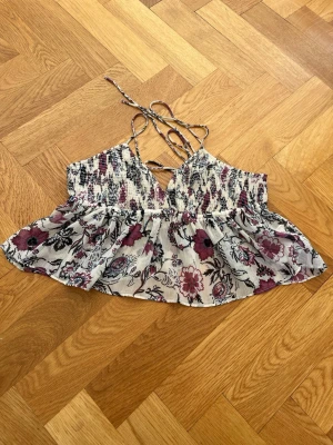 Blommig linnetopp med smock och knyt - Supersöt blommig linnetopp med smock upptill och tunna axelband som knyts i ryggen. Toppen har volang nedtill och ett luftigt, flowy tyg i vitt med lila och svarta blommor. Perfekt för varma dagar och ger en romantisk vibe.