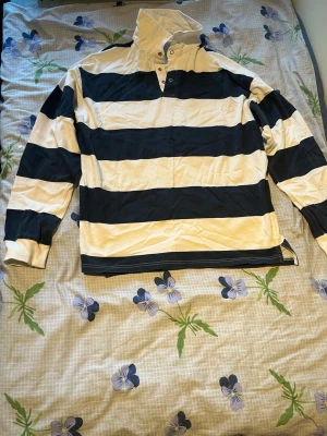 Randig rugbytröja med hög krage - Snygg rugbytröja med breda marinblå och vita ränder, hög krage och knappar vid halsen. Tröjan har långa ärmar och en avslappnad passform, perfekt för en sportig och casual stil. Materialet känns mjukt och bekvämt.