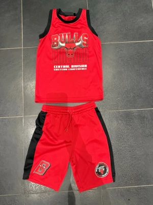 Chicago Bulls basketset NBA - Rött basketset med Chicago Bulls-logga, består av ärmlös tröja och shorts med svarta detaljer på sidorna. Materialet är lätt och andas, perfekt för basketträning eller match. Märkt med NBA och Bulls, passar för ungdomar ca 9-10 år.