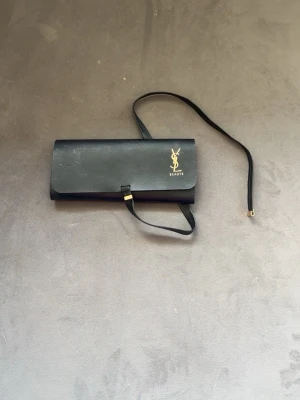 Svart kuvertväska från Yves Saint Laurent - Stilren svart kuvertväska från Yves Saint Laurent med guldigt YSL-logo och texten 'BEAUTÉ' på framsidan. Väskan har en smal axelrem och är tillverkad i slätt material med enkel, rektangulär form. Perfekt för dig som gillar klassisk och elegant design.