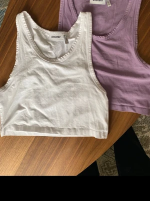 2 croppade linnetoppar Weekday - Två croppade linnetoppar från Weekday, en i vitt och en i ljuslila. Båda är ärmlösa och har en enkel, stilren design. Perfekta för varma dagar eller som lager under skjorta eller jacka.