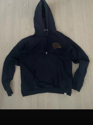Marinblå les deux hoodie med tryck - Snygg marinblå hoodie med stor huva och dragsko. Framtill finns en känguruficka och ett gult tryck på bröstet. Baksidan har ett stort gult tryck med texten 'Harajuku Teahouse & Boardshop' och illustration av ett tehus. Perfekt för en avslappnad streetwear-look.