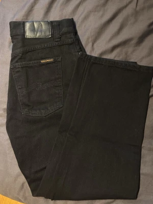 Svarta wide jeans från Nudie Jeans - Säljer ett par svarta jeans från Nudie Jeans med klassisk femficksdesign och diskret logga på bakfickan. Jeansen har raka ben och är tillverkade i ett mjukt bomullstyg som sitter snyggt. Perfekta för dig som gillar stilrena och tidlösa plagg.