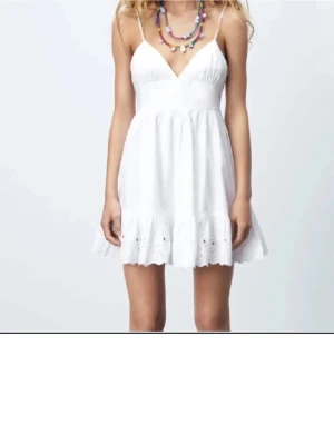 Vit kort sommarklänning med spets - Söt vit kortklänning med tunna axelband och v-ringning. Kjolen har volangkant med broderad spetsdetalj som ger en romantisk känsla. Perfekt för varma dagar och sommarhäng. Tillverkad i lätt bomullstyg som känns luftigt och bekvämt.