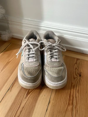 Nike Air Force 1 Shadow beige pastell - Säljer ett par Nike Air Force 1 Shadow sneakers i beige med detaljer i pastellgrönt och ljusrosa. Skorna har klassisk låg siluett, perforerad tå och dubbla lager på swoosh och häl. Tillverkade i läder och textil med vit sula och snörning.