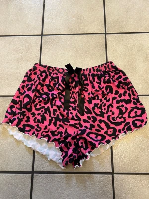 Rosa leopardmönstrade pyjamasshorts - Snygga pyjamasshorts i rosa med svart leopardmönster och vit volangkant. Shortsen har en svart rosett framtill och elastisk midja för extra komfort. Perfekta för chillkvällar hemma.