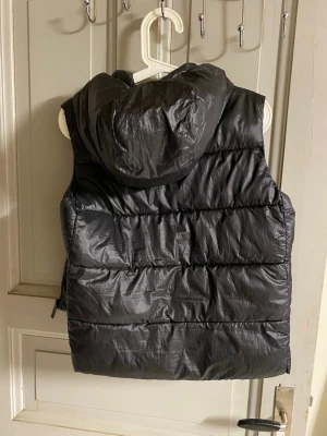 Svart dunväst med huva - Snygg svart dunväst med huva och quiltad design. Västen har en glansig finish och breda kanaler som ger en modern look. Perfekt att slänga över en hoodie eller tröja för extra värme. Passar dig som gillar streetwear och enkel stil.