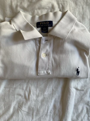 Vit pikétröja från Polo Ralph Lauren - Klassisk vit pikétröja från Polo Ralph Lauren med korta ärmar, krage och knappar framtill. Tröjan har den ikoniska broderade loggan i marinblått på bröstet och är tillverkad i mjuk bomull. Perfekt basic-plagg för en clean och stilren look. Skriv vid minsta fundering det är barn storlek 14-16 år