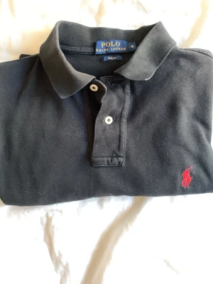Svart pikéskjorta från Polo Ralph Lauren - Svart pikéskjorta från Polo Ralph Lauren med klassisk krage, två knappar framtill och röd broderad logga på bröstet. Tillverkad i mjuk bomull och har korta ärmar för en avslappnad men stilren look. Skriv vid minsta fundering
