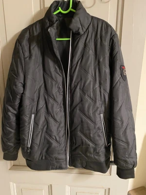 Svart quiltad pufferjacka - Snygg svart pufferjacka med quiltat mönster och ståkrage. Jackan har två fickor med dragkedja och ribbade muddar vid ärmslut och nederkant. Märke på vänster ärm. Perfekt för kalla vinterdagar och riktigt skön att bära.