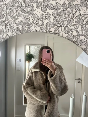 Beige teddyjacka med dragkedja - Mysig beige teddyjacka med hög krage och dragkedja framtill. Jackan har en oversized passform och stora fickor på sidorna. Perfekt för kalla dagar när du vill vara både varm och trendig.