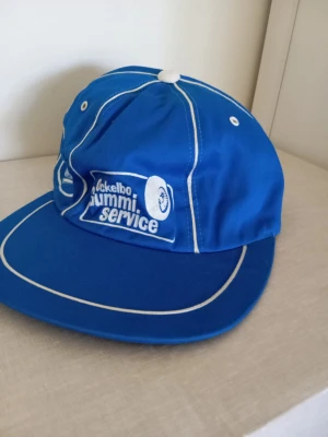 Blå keps från Ockelbo Gummi Service - Snygg blå keps med vit piping och broderad logga från Ockelbo Gummi Service framtill. Klassisk modell med böjd skärm och ventilationshål. Tillverkad i slitstarkt tyg, perfekt för dig som gillar retrostil och unika accessoarer.