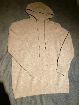 Beige stickad hoodie med huva - Mysig beige stickad hoodie med dragsko i huvan och ribbade muddar. Perfekt för dig som gillar en enkel och stilren look. Materialet känns mjukt och passar bra till många outfits.