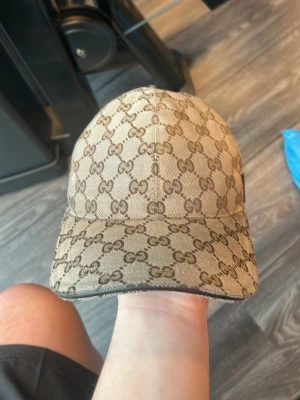 Gucci keps med monogrammönster - Snygg beige keps från Gucci med klassiskt GG-monogram i brunt över hela kepsen. Justerbar rem baktill och böjd skärm. Perfekt accessoar för att lyfta din outfit med en lyxig touch.