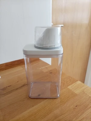 Förvaringsburk med måttbägare - Transparent förvaringsburk i plast med vitt lock och tillhörande måttbägare på toppen. Måttbägaren har tydliga röda markeringar för ml och cups, perfekt för att mäta upp tvättmedel. Finns nån tejp på den ja int tagit bort men går bort om man vill