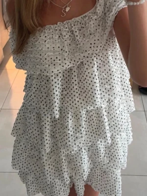 Vit prickig volangklänning - Söt vit kortklänning med svarta prickar och flera lager volanger. Offshoulder-modell som ger en romantisk vibe. Perfekt för sommardagar och har en luftig passform. Tillverkad i lätt och bekvämt material.