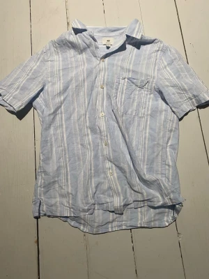 Blårandig kortärmad skjorta H&M - Snygg kortärmad skjorta från H&M i ljusblått med vita vertikala ränder. Skjortan har klassisk krage, bröstficka och knäppning framtill. Tillverkad i ett lätt och luftigt tyg, perfekt för varma dagar.