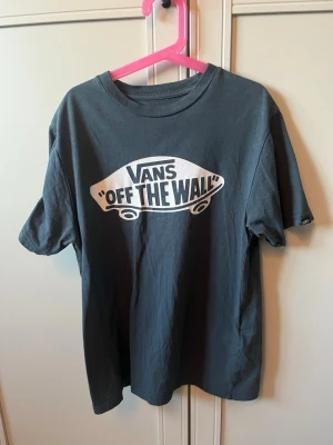 Grön Vans Off The Wall t-shirt - Snygg grön t-shirt från Vans med det klassiska 'Off The Wall'-trycket i vitt framtill. Tillverkad i 100% bomull för en skön och avslappnad känsla. Perfekt för dig som gillar streetwear och skatekultur.