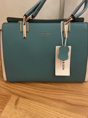 Blågrön handväska från Jones & Son - Snygg handväska i blågrönt konstläder från Jones & Son. Väskan har guldfärgade metalldetaljer, dubbla handtag och vita paneler på sidorna. Stilren och boxig form som ger en modern look. Perfekt för dig som vill ha en trendig accessoar till din outfit.