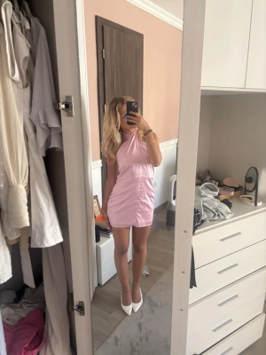 Rosa omlott kortklänning i satin - Snygg rosa kortklänning i satin med omlottdesign och halterneck. Klänningen har en figurnära passform och slät, glansig yta som ger en lyxig känsla. Perfekt för dig som vill sticka ut med en feminin och trendig look.