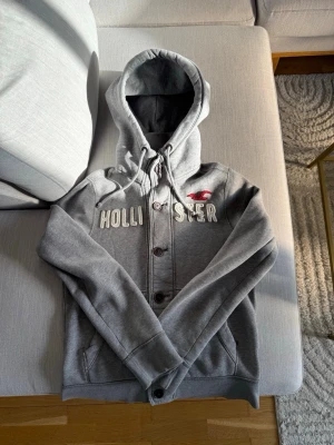 Grå hoodie från Hollister med knappar - Snygg grå hoodie från Hollister med broderad logga och text på bröstet. Hoodien har knappar framtill, dragsko i huvan och två fickor. Insidan är mjuk och fleecefodrad för extra komfort. Perfekt för chill dagar och streetwear vibes. Priset går att diskutera om