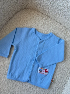 Ljusblå babykofta med bilar - Supersöt ljusblå babykofta från Spring Garden med tryckknappar framtill och en liten ficka med broderade bilar i flera färger. Tillverkad i mjukt ribbat tyg, perfekt för små barn. Koftan har långa ärmar och rund hals. 0-3månader. Superfint skick! 💛