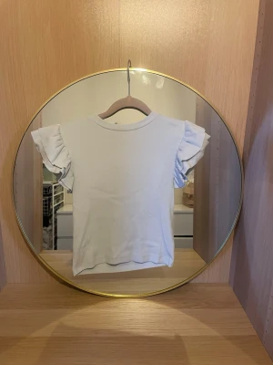 Vit topp med volangärm från Gina Tricot - Söt vit topp från Gina Tricot med korta volangärmar som ger en extra touch till outfiten. Tillverkad i mjuk bomull för skön känsla och enkel att matcha med olika stilar. Perfekt för dig som gillar en enkel men ändå trendig look.skriv för fler bilder💗pris kan diskuteras!skulle säga att den sitter som en xxs/xs💕