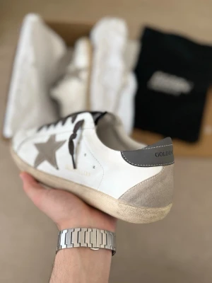 Golden Goose Superstar vita/grå sneakers - Säljer ett par stilrena Golden Goose sneakers i vit/grå färg. Skorna är använda cirka 10 gånger och är i väldigt bra skick överlag. Enda anmärkningen är att skosnörena har fått en färgförändring efter kontakt med klor, vilket gör att de skiljer sig något i färg. Skosnören är enkla att byta ut om man vill.