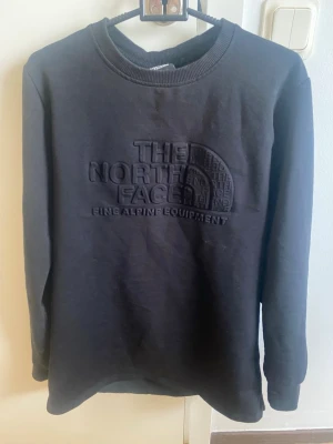 Svart sweatshirt från The North Face - Säljer en svart sweatshirt från The North Face med upphöjt logotryck på bröstet. Tröjan har rund halsringning och långa ärmar. Perfekt för dig som gillar stilrena och bekväma plagg med en sportig touch.