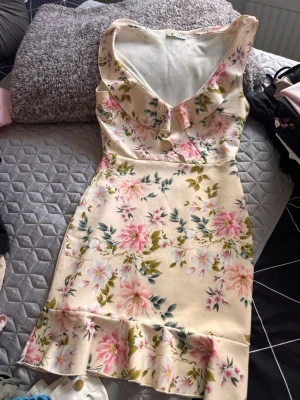 Blommig kortklänning med volang - Söt kortklänning i ljusbeige med rosa och grönt blommönster. Klänningen har v-ringning, volangdetaljer vid halsringningen och nederkanten samt är ärmlös. Perfekt för sommarens alla tillfällen.