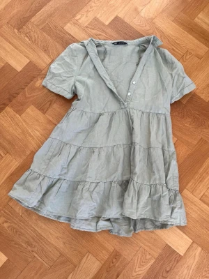 Ljusgrön volangklänning med knappar - Söt ljusgrön kortklänning med volanger och korta ärmar. Klänningen har v-ringning med knappar upptill och är tillverkad i ett lätt bomullstyg. Perfekt för varma dagar och enkel att styla med sneakers eller sandaler.