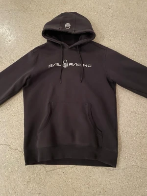 Svart hoodie från Sail Racing - Svart hoodie från Sail Racing med vit logga och text på bröstet. Klassisk känguruficka framtill och justerbar huva med dragsko. Mjuk insida och ribbade muddar vid ärmar och nederkant. Perfekt för en avslappnad och sportig stil.