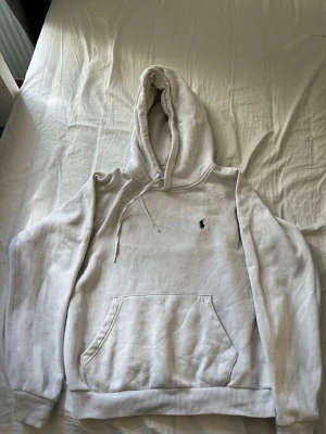 Vit hoodie från Polo Ralph Lauren - Snygg vit hoodie från Polo Ralph Lauren med klassisk huva och dragsko. Har en känguruficka framtill och den ikoniska lilla blå loggan broderad på bröstet. Tillverkad i mjukt material som känns skönt mot huden. Perfekt för en chill och stilren look. Den har inga hål men den har bara massa fläckar som inte går bort i tvätten