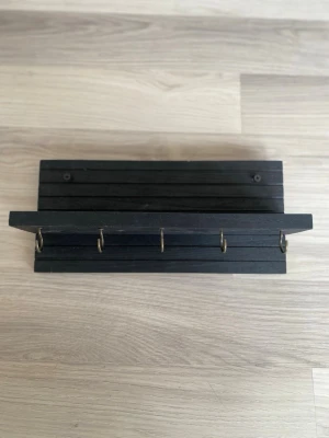 Svart nyckelhängare i trä - Snygg svart nyckelhängare i trä med fem metallkrokar. Perfekt för att hålla ordning på nycklar eller småsaker i hallen. Enkel och stilren design som passar de flesta inredningsstilar.
