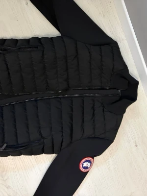 Svart cardigan från Canada Goose - Snygg svart cardigan från Canada Goose med quiltad front och ärmar i stretchigt material. Jackan har dragkedja framtill, två sidofickor med dragkedja och den klassiska Canada Goose-loggan på ärmen. Perfekt för kalla vinterdagar.