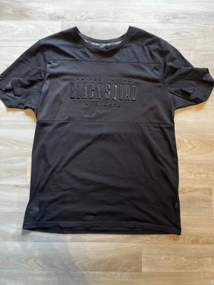 Svart Black Squad t-shirt - Snygg svart t-shirt från Black Squad med upphöjt tryck på bröstet där det står 'Raised in the Black Squad Projects'. Klassisk rund halsringning och korta ärmar. Perfekt för en clean och modern stil.