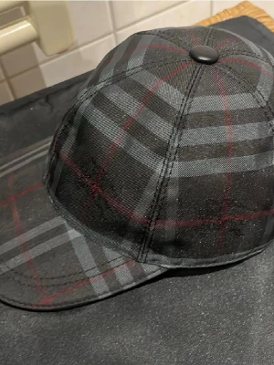 Burberry keps - Snygg keps i svart med rutigt mönster i grått och tunna röda linjer. Klassisk böjd skärm och knapp på toppen. Tillverkad i ett mjukt tygmaterial som känns bekvämt på huvudet. Perfekt accessoar för en stilren och trendig look.