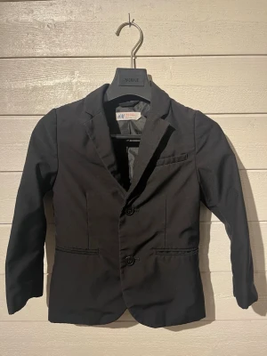 Svart kavaj från H&M barn - Stilren svart kavaj för barn från H&M med två knappar framtill, klassisk krage och fickdetaljer. Perfekt för dig som vill ha en enkel och snygg look. Tillverkad i ett mjukt tyg med slät finish och helfodrad insida. 4-5 år.