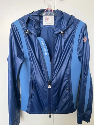 Blå vindjacka från Moncler med huva - Snygg blå vindjacka från Moncler med två nyanser av blått, huva med dragsko och dragkedja framtill. Jackan har Moncler-logga på ärmen, smal passform och sidofickor med knappar. Materialet är glansigt och lätt, perfekt för blåsiga dagar.