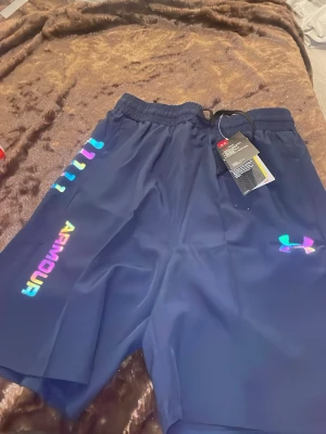 Blå träningsshorts från Under Armour - Snygga marinblå träningsshorts från Under Armour med regnbågsfärgad logga och text på sidan. Shortsen har elastisk midja och är tillverkade i ett lätt och snabbtorkande syntetmaterial, perfekta för gymmet eller löprundan.
